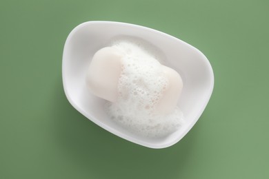 Photo of Dish with soap bar and fluffy foam on green background, top view