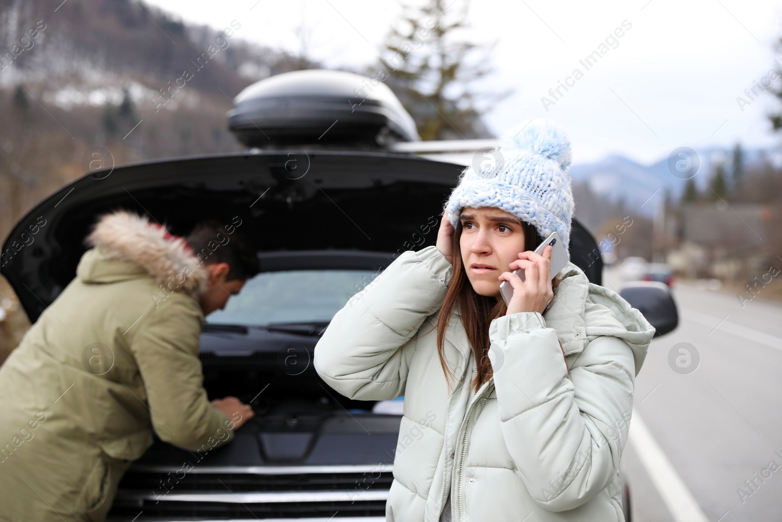 Photo of Emotional young woman talking on phone while stressed man repairing broken car outdoors in winter