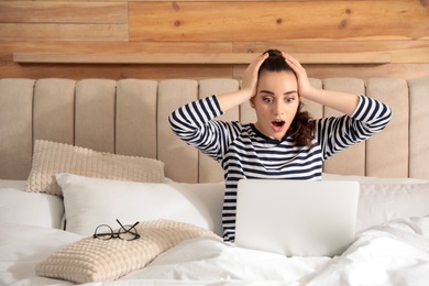Photo of Shocked young woman working with laptop on bed at home