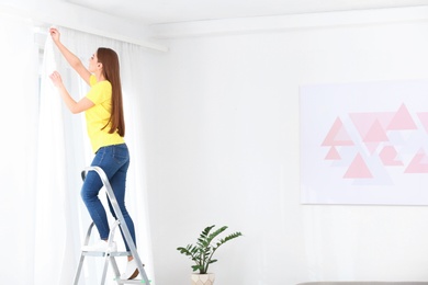 Photo of Young woman hanging window curtain in room. Space for text