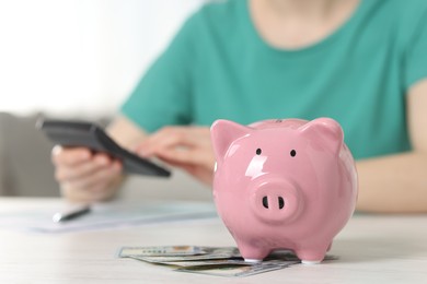 Financial savings. Woman using calculator at white wooden table indoors, focus on piggy bank and dollar banknotes