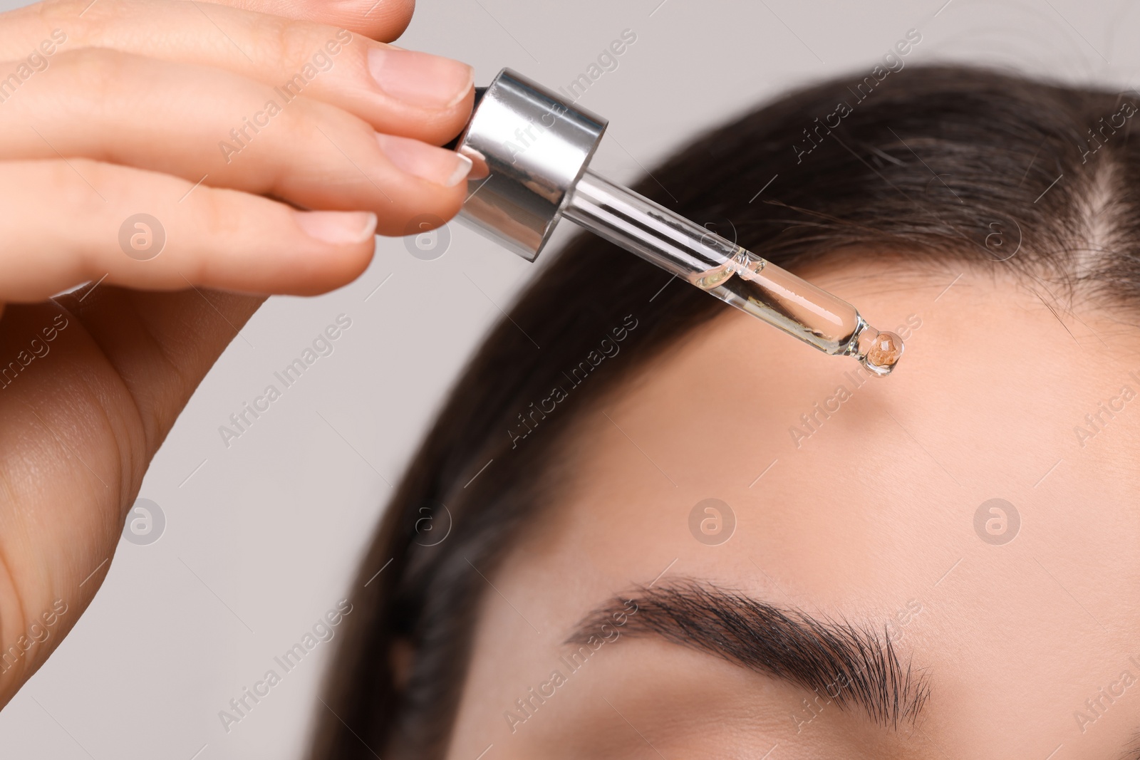 Photo of Young woman applying essential oil onto face on light grey background, closeup