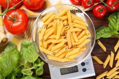 Photo of Kitchen scale with bowl of pasta among tomatoes and basil on wooden table, flat lay