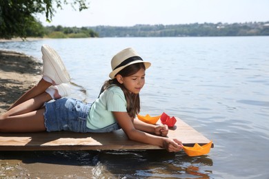 Photo of Cute little girl playing with paper boat on wooden pier near river