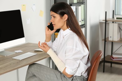 Photo of Young woman with arm wrapped in medical bandage talking on phone at workplace