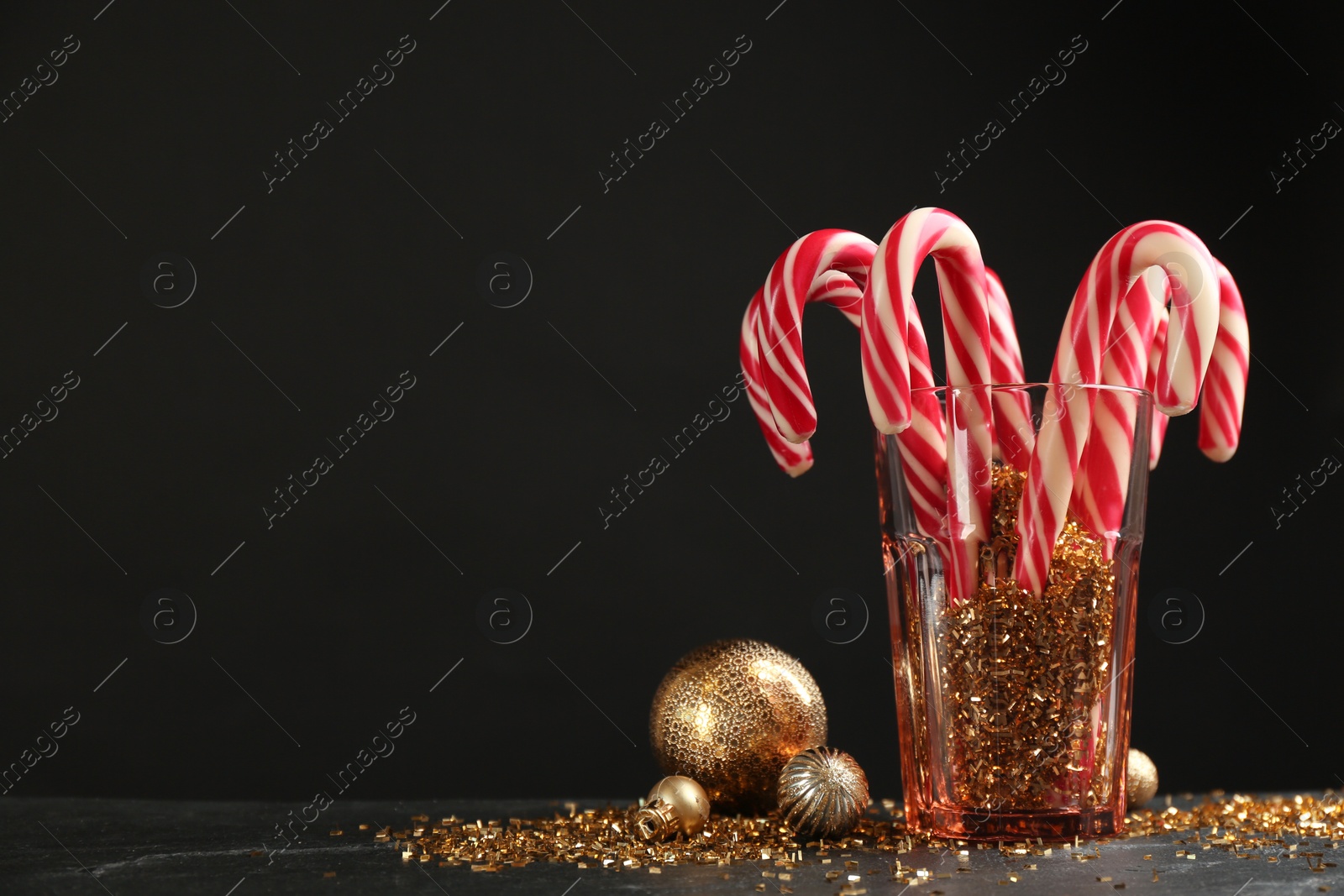 Photo of Candy canes in glass and Christmas balls on black table against dark background, space for text