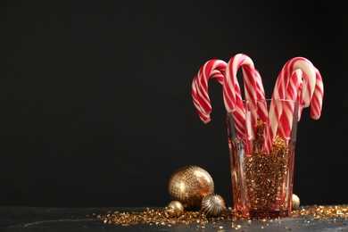 Photo of Candy canes in glass and Christmas balls on black table against dark background, space for text