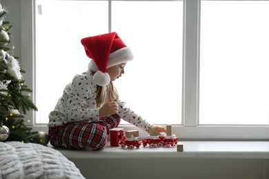 Photo of Cute little girl in Santa hat playing with Christmas candleholder on window sill at home