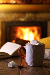 Photo of Mug with hot cocoa, marshmallows and book on wooden table near fireplace