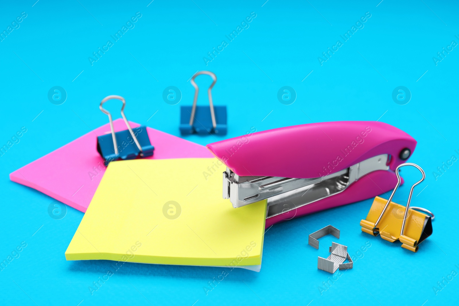 Photo of New bright stapler with blinder clips and sticky notes on light blue background. School stationery