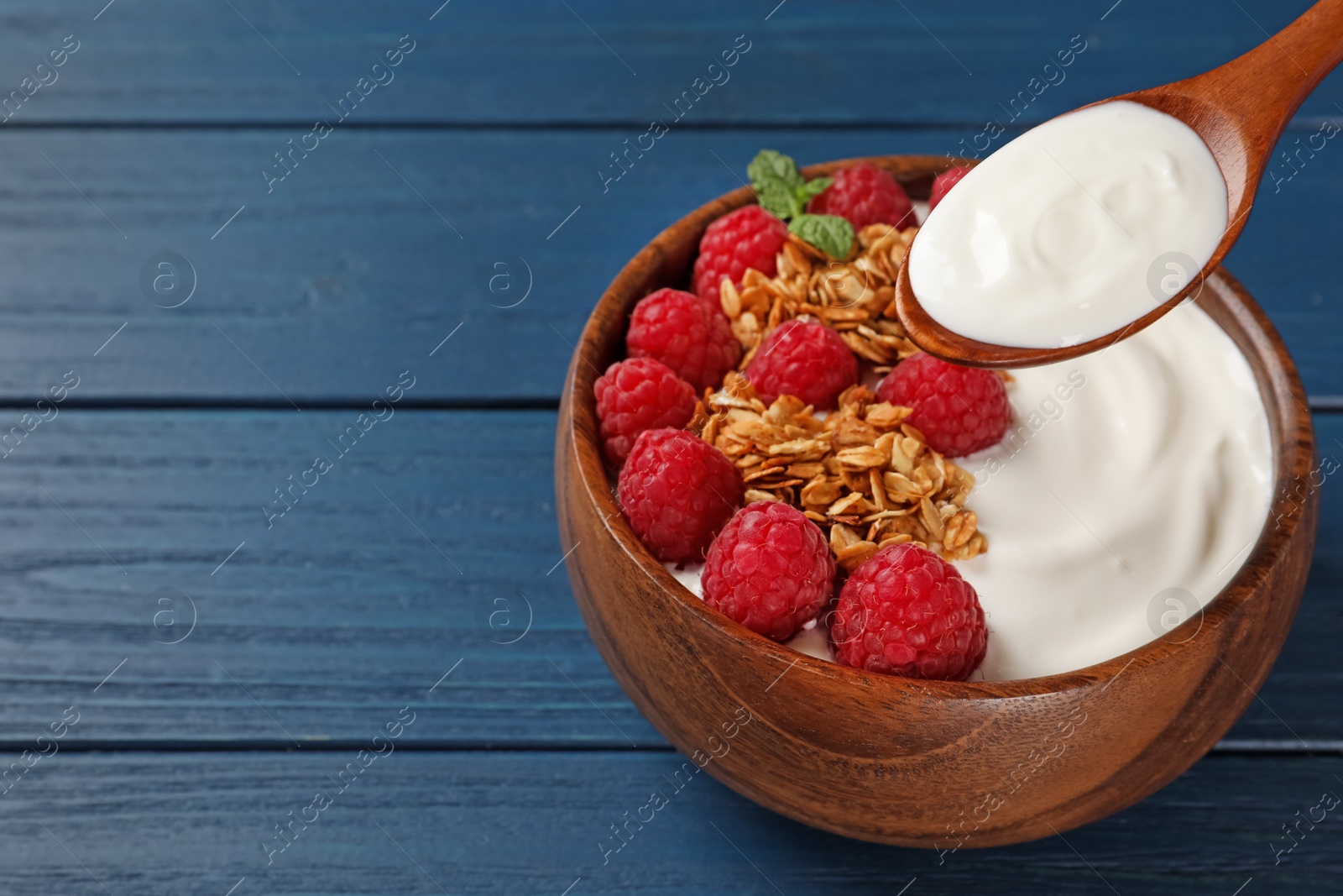 Photo of Eating tasty yogurt with raspberries and muesli from bowl on blue wooden table. Space for text