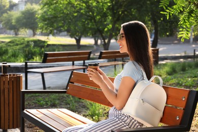 Beautiful young woman with stylish backpack and cup of coffee on bench in park