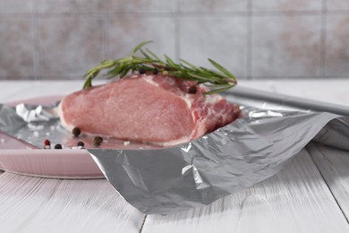 Photo of Aluminum foil with raw meat, rosemary and spices on white wooden table
