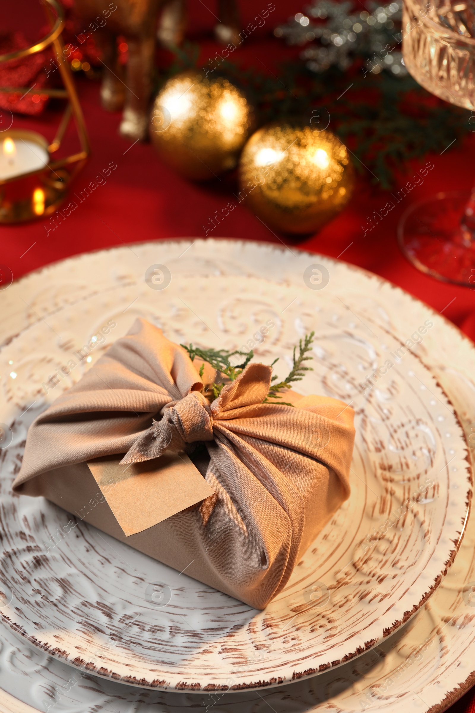 Photo of Furoshiki technique. Gift packed in beige fabric, blank card and festive decor on red table