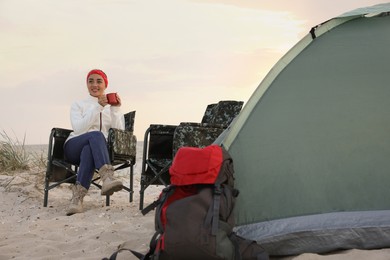Photo of Young woman with cup of hot drink near camping tent on sandy beach