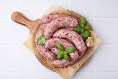 Photo of Board with raw homemade sausages, basil leaves and peppercorns on white tiled table, top view