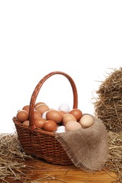 Wicker basket full of chicken eggs and dried straw on wooden table against white background