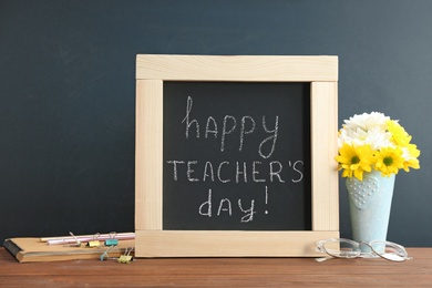 Photo of Little blackboard with inscription HAPPY TEACHER'S DAY, flowers and stationery on table