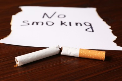 Broken cigarette and words No Smoking written on paper on wooden table, closeup