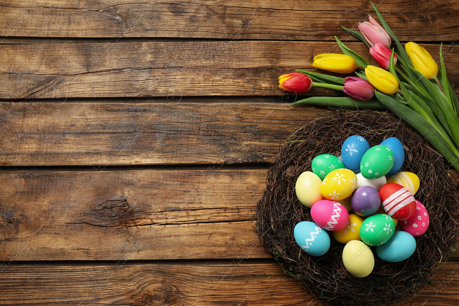 Photo of Colorful Easter eggs in decorative nest and tulips on wooden background, flat lay. Space for text