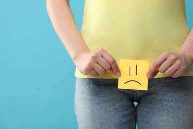 Cystitis. Woman holding sticky note with drawn sad face on light blue background, closeup. Space for text