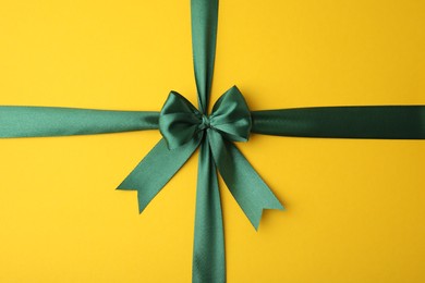 Green satin ribbon with bow on yellow background, top view