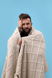 Photo of Man wrapped in blanket suffering from headache on light blue background. Cold symptoms
