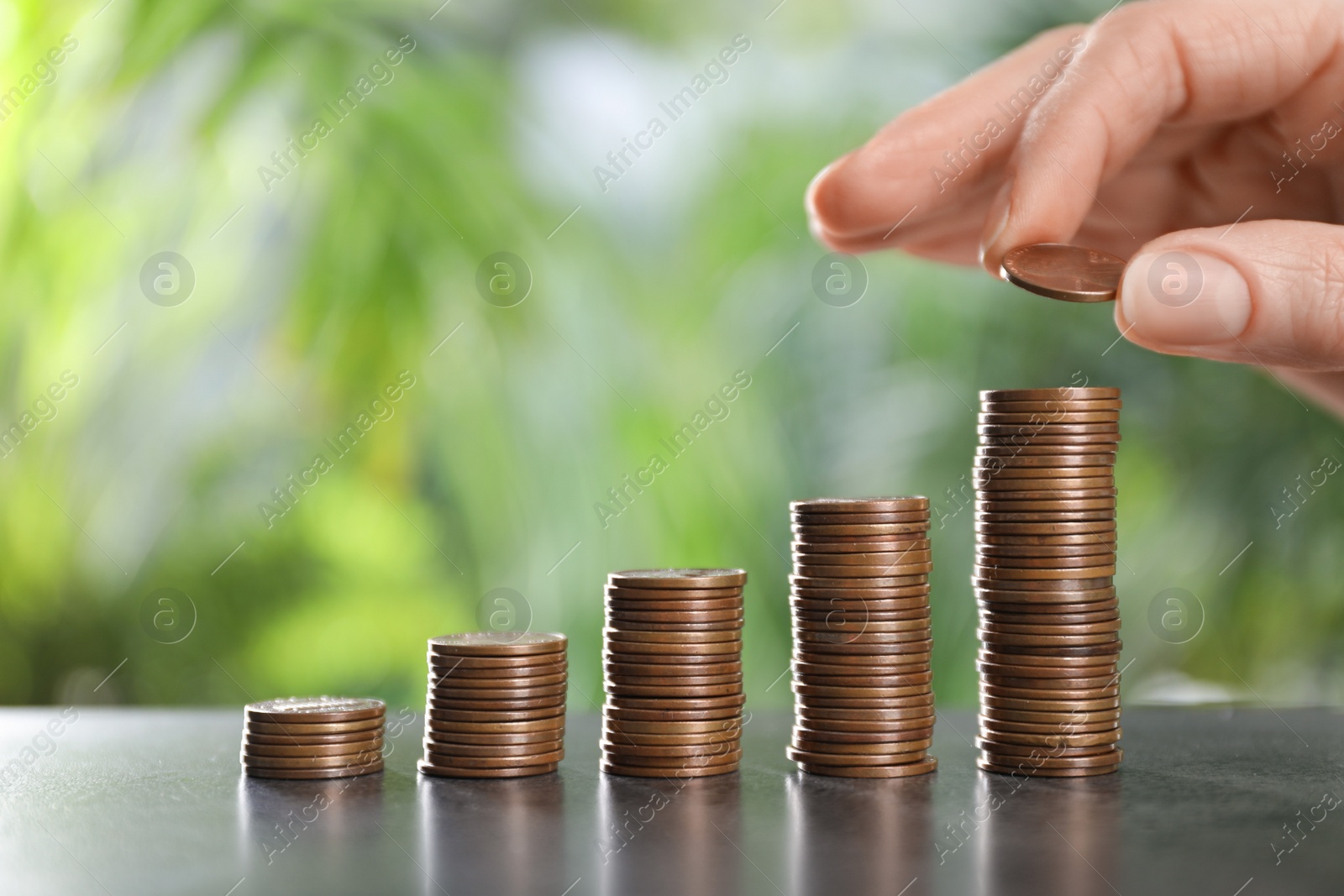 Photo of Woman stacking coins at grey table against blurred green background, closeup. Space for text