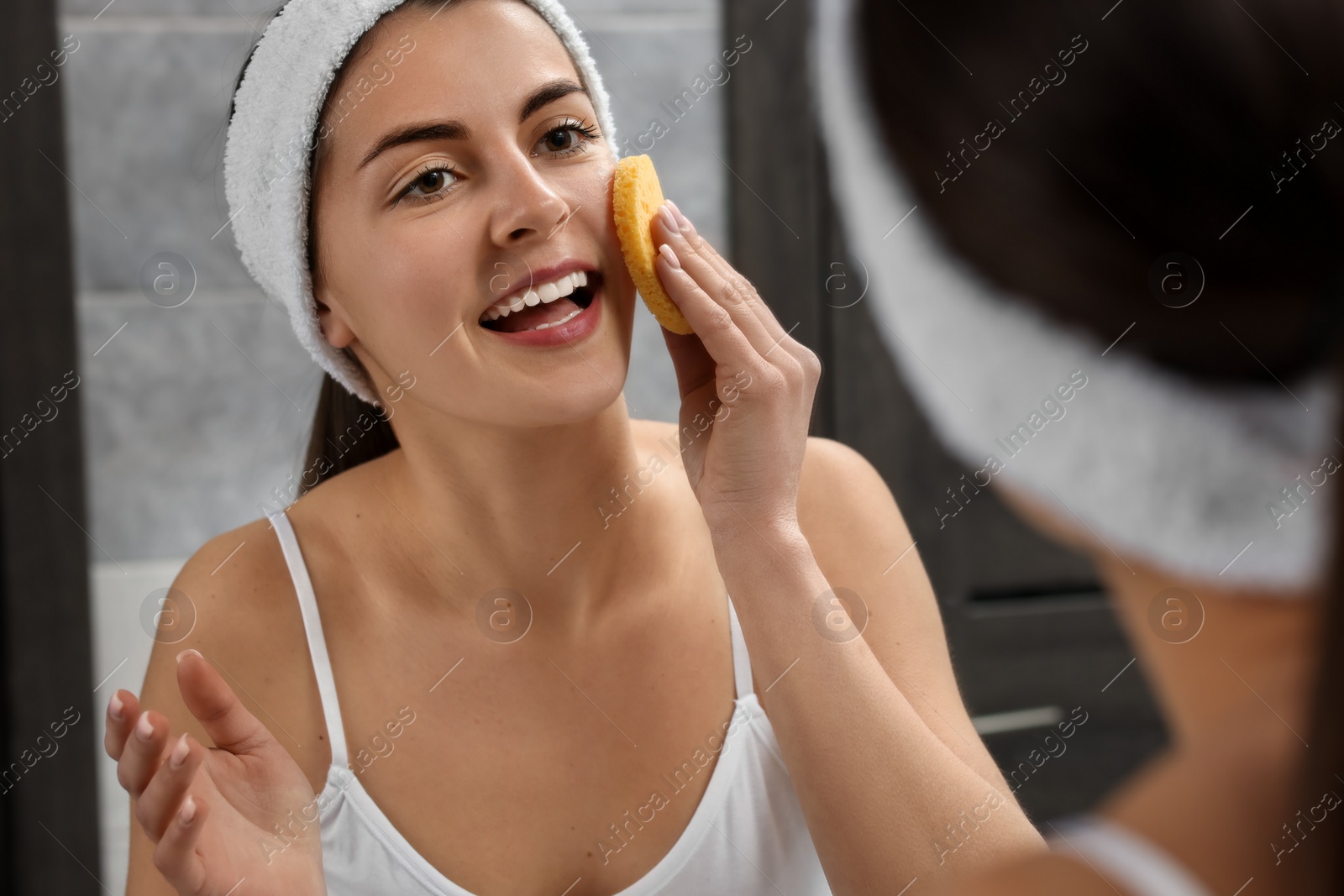 Photo of Young woman with headband washing her face using sponge near mirror in bathroom
