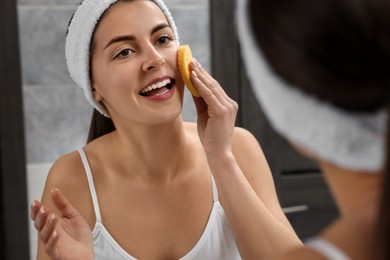 Young woman with headband washing her face using sponge near mirror in bathroom