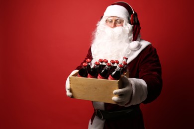 Photo of MYKOLAIV, UKRAINE - JANUARY 18, 2021: Santa Claus with headphones holding wooden crate full of Coca-Cola bottles on red background