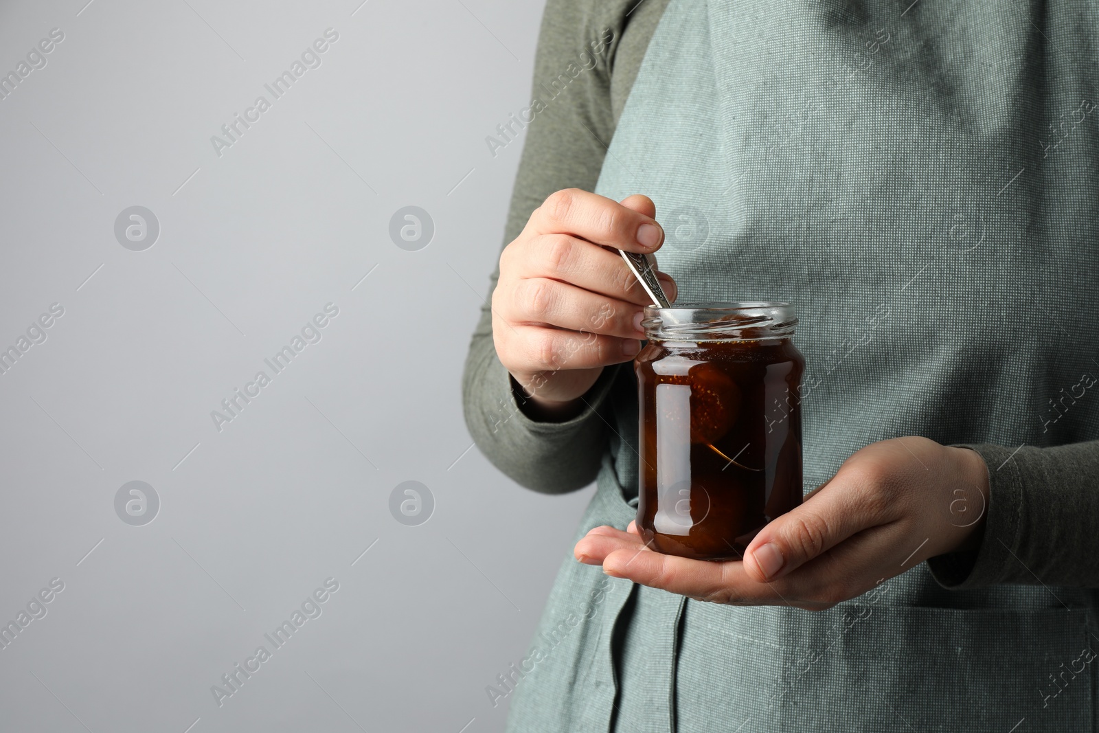 Photo of Woman holding jar of tasty sweet fig jam on grey background, closeup. Space for text