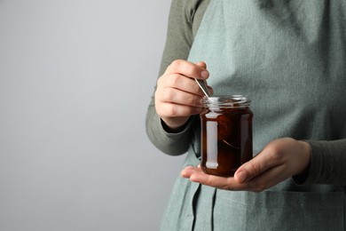 Photo of Woman holding jar of tasty sweet fig jam on grey background, closeup. Space for text