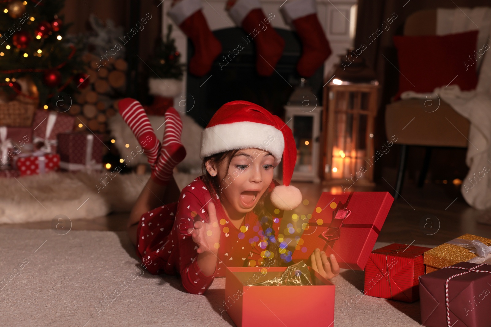 Photo of Surprised child in Santa hat opening Christmas gift on floor at home