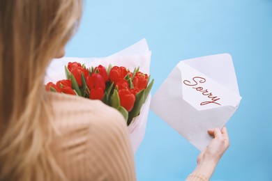 Image of Apology. Woman holding Sorry greeting card and bouquet of tulips on light blue background, closeup