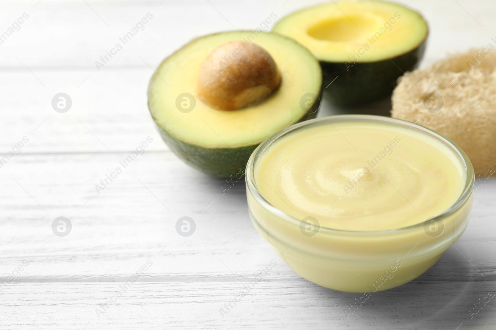 Photo of Handmade face mask in glass bowl and avocado on white wooden table