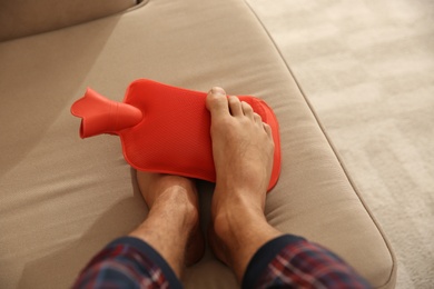 Photo of Man warming feet with hot water bottle on sofa, closeup