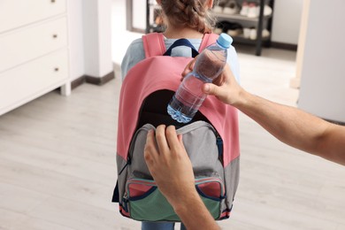 Photo of Father putting bottle of water into daughter`s backpack at home, closeup. Preparing to school