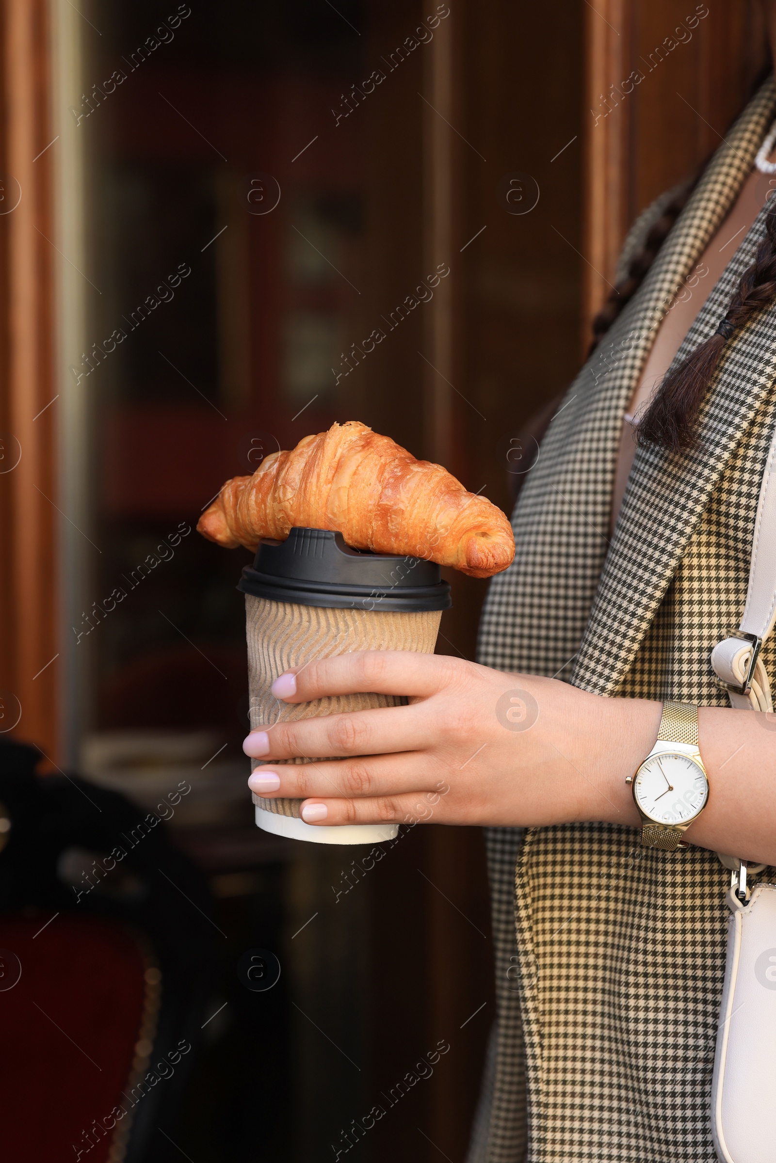 Photo of Woman holding tasty croissant and cup of coffee near wooden door outdoors, closeup