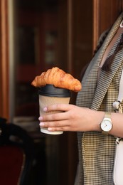 Woman holding tasty croissant and cup of coffee near wooden door outdoors, closeup