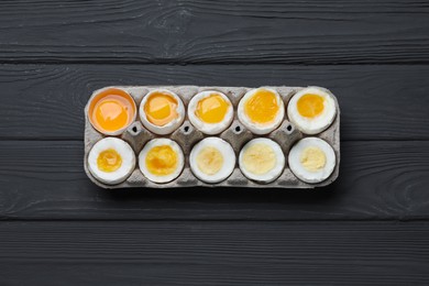 Boiled chicken eggs of different readiness stages in carton on black wooden table, top view