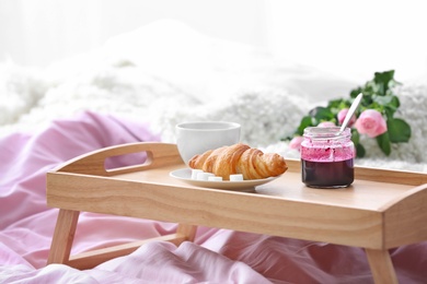 Photo of Wooden tray with tasty croissant, cup of coffee and jam on bed