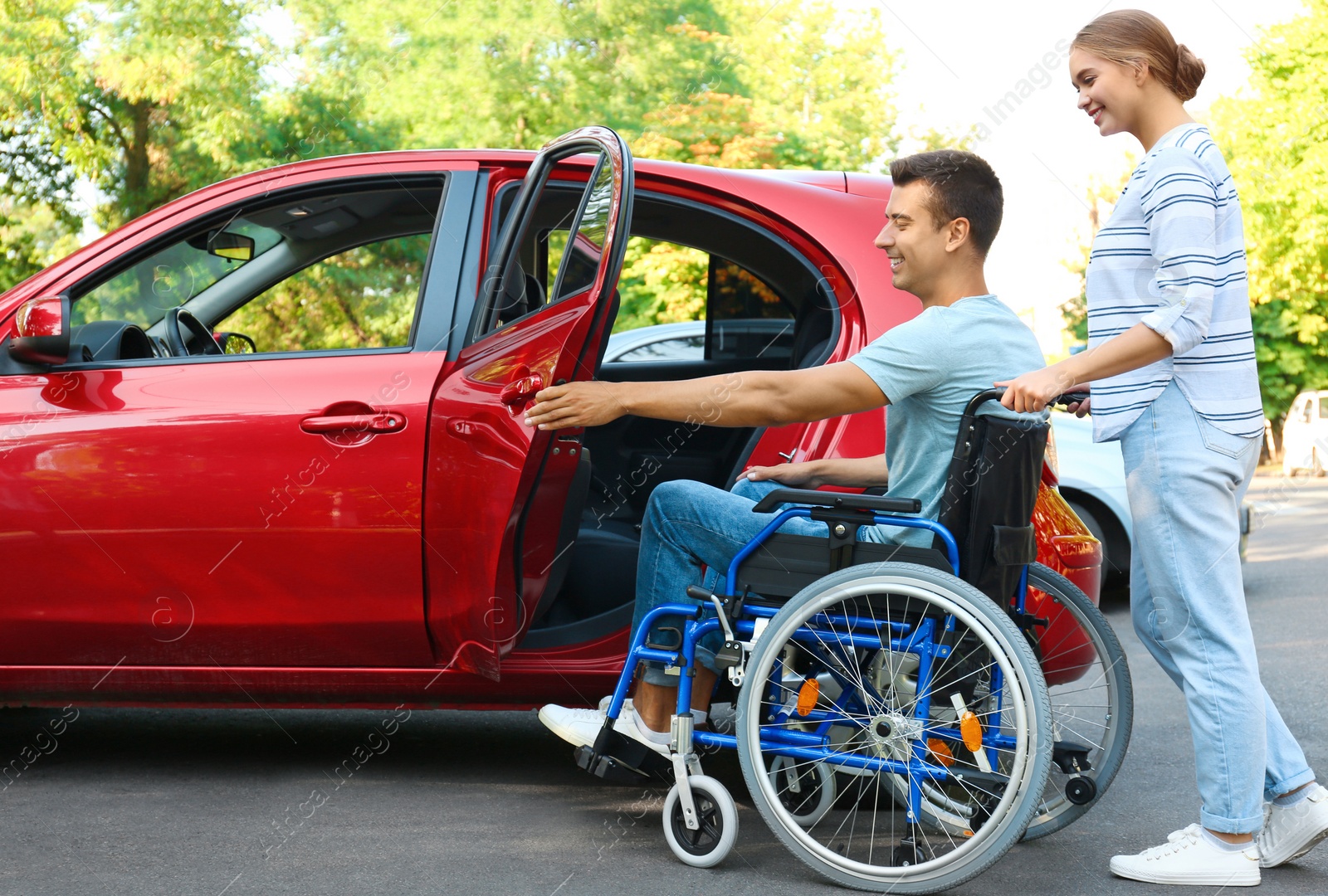 Photo of Young woman helping disabled man in wheelchair to get into car outdoors