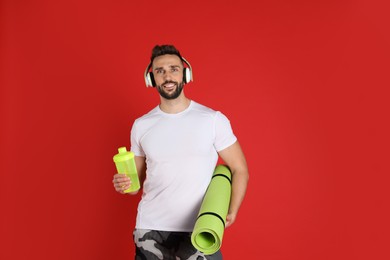 Photo of Handsome man with headphones,yoga mat and shaker on red background