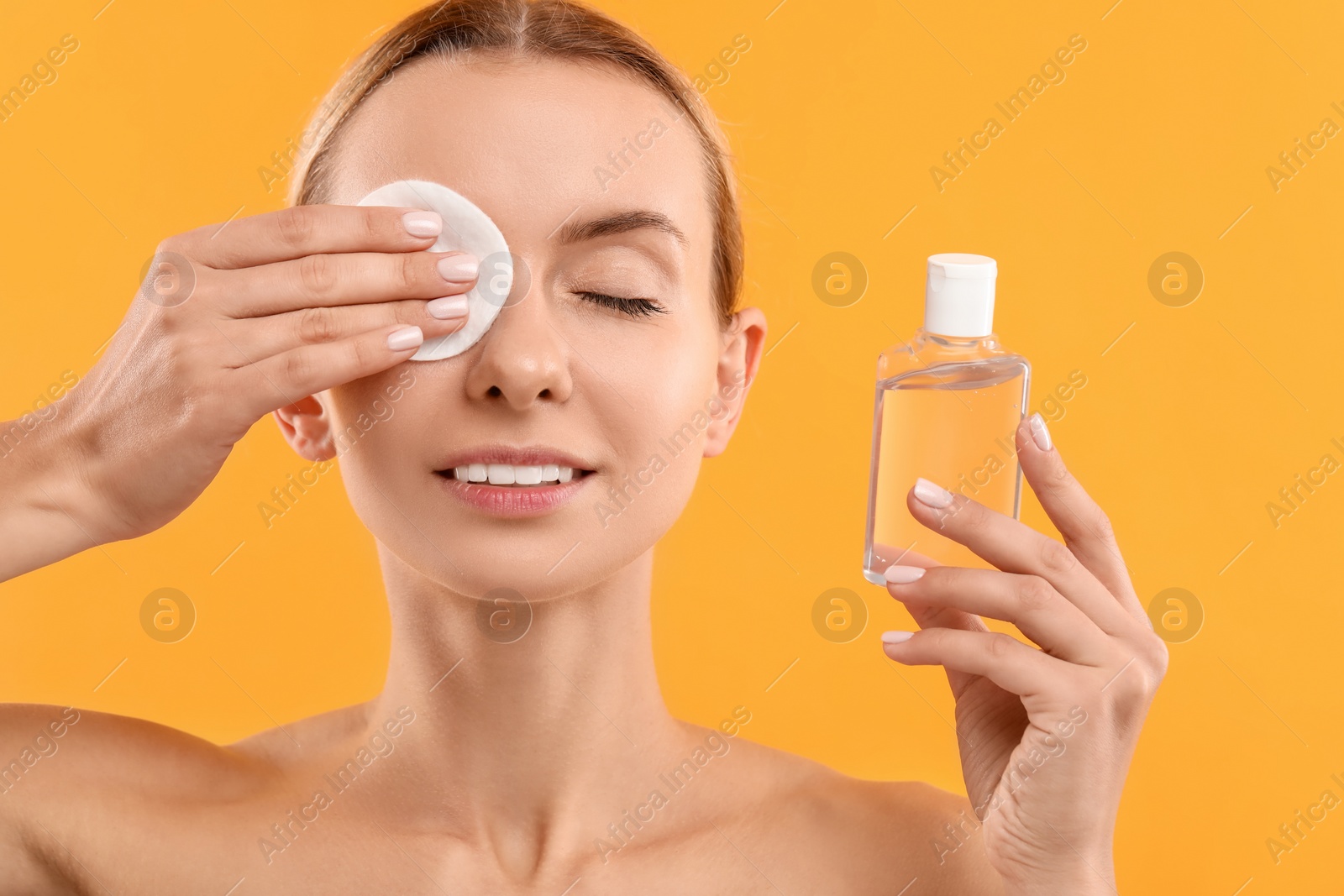 Photo of Beautiful woman removing makeup with cotton pad and holding bottle on yellow background, closeup