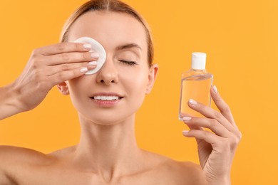 Photo of Beautiful woman removing makeup with cotton pad and holding bottle on yellow background, closeup