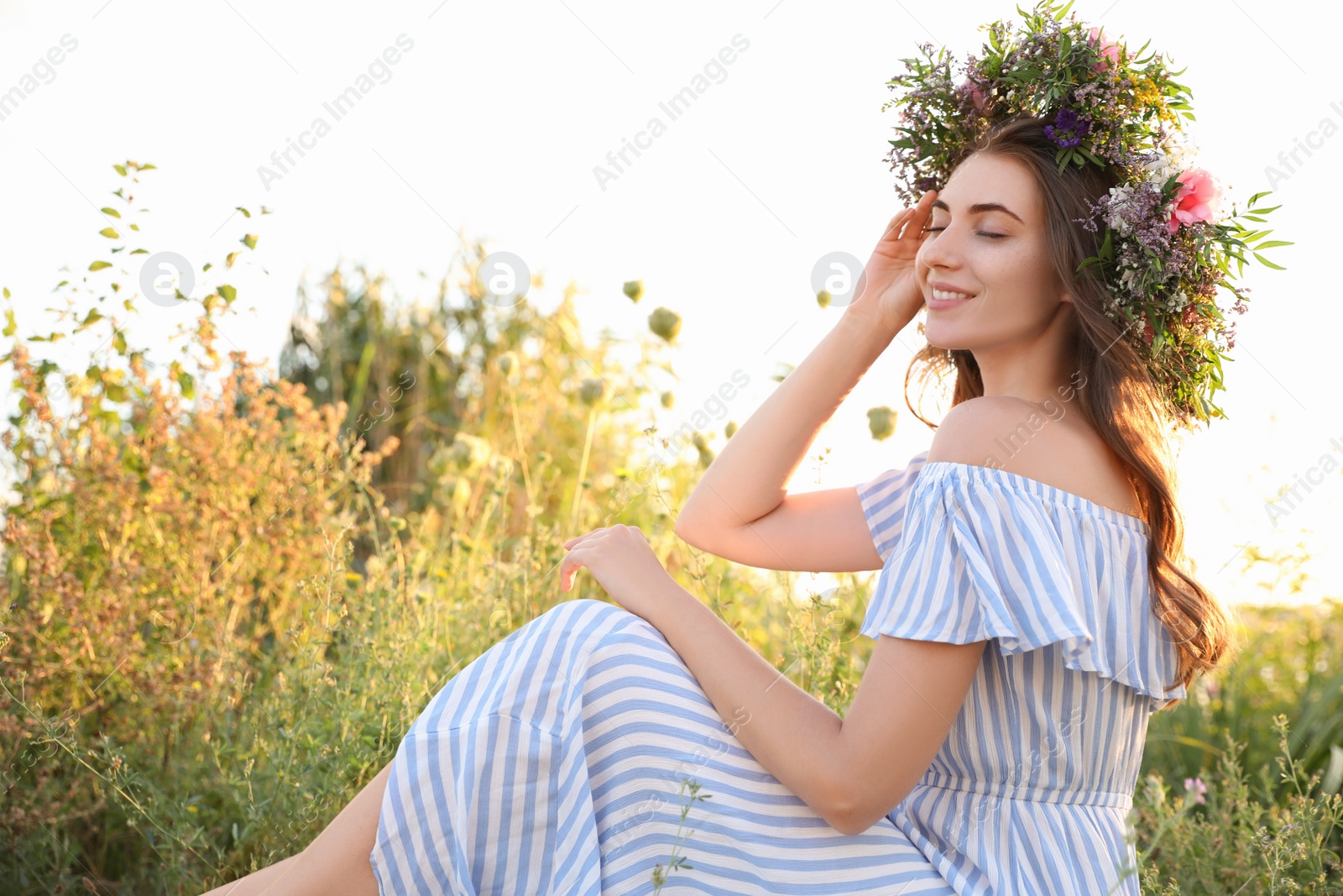 Photo of Young woman wearing wreath made of beautiful flowers outdoors on sunny day