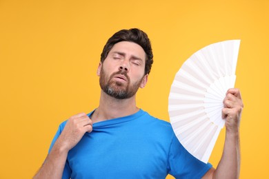 Photo of Unhappy man with hand fan suffering from heat on orange background