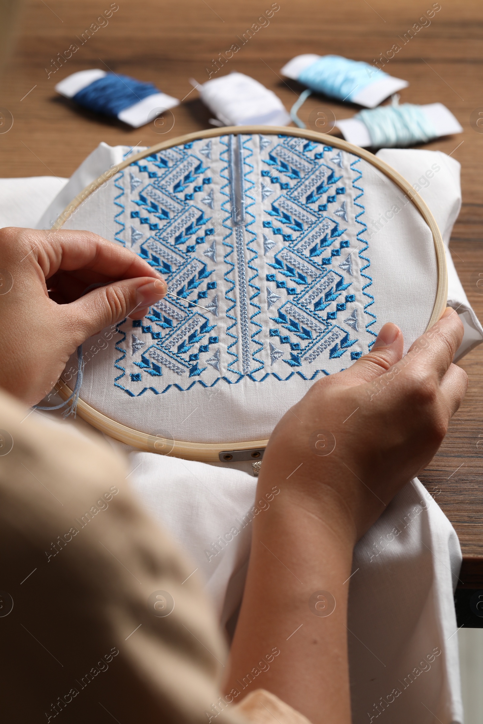 Photo of Woman embroidering white shirt with blue thread at wooden table, closeup. Ukrainian national clothes