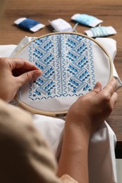 Woman embroidering white shirt with blue thread at wooden table, closeup. Ukrainian national clothes